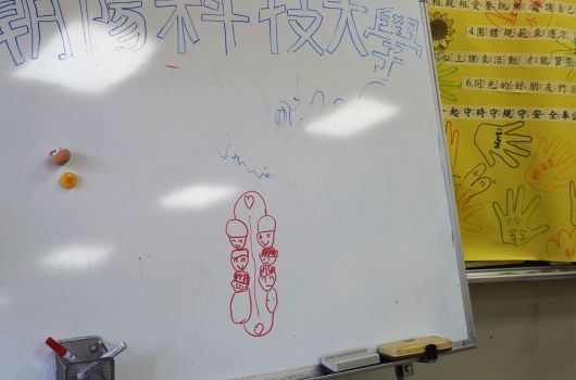 教室白板上用紅色筆畫著兩個手掌圖案，板上方有中文標題。左側放置兩顆球和紅色物品，右側貼著黃色安全宣傳海報。板面下方有粉筆擦。
