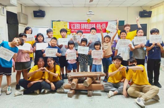 一群小學生在教室內進行活動，穿著黃色和其他顏色衣服的學生舉著證書，面帶笑容。前排學生圍坐成圈，後排學生站立舉著獎狀。教室牆上掛著紅色的活動布幕，氣氛歡樂。