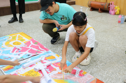 兩位年輕人在室內地板上進行美勞活動，正在製作彩色紙質手工藝品。他們蹲坐在鋪展的繽紛色紙旁，認真地進行剪裁和組合工作。背景可見室內裝飾和其他玩具用品。