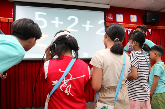 教室裡，一群學童面向投影螢幕觀看數學題目「5+2+1」。講台上方掛著紅色布幔和中文標誌，學生穿著校服認真聽講，呈現課堂教學情景。