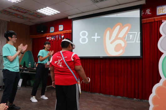 室內教室進行數學教學活動，講師在投影幕前展示「8+6=」的數學題目，搭配手勢示意。穿著紅色和青綠色T恤的參與者站在講師周圍，認真觀看投影幕上的數學問題。背景有紅色布幕和教室設施。