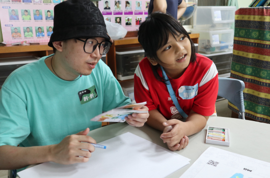 兩位學生在教室內坐在桌前進行美術創作。左邊穿著綠色上衣、戴黑色帽子和眼鏡的學生，與右邊穿著紅色上衣的學生一起使用彩色筆在白紙上作畫。背景可見書架、教室布告欄和學生作品展示。