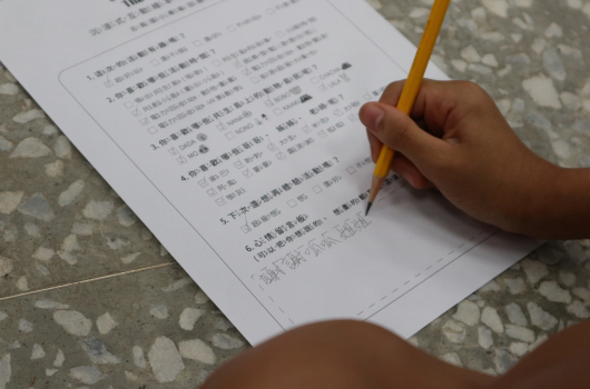 一名學生手握黃色鉛筆，在印有中文文字的試卷或作業紙上作答。紙張放在有花紋的灰白色地面上，學生正專注地進行書寫或標記答案。