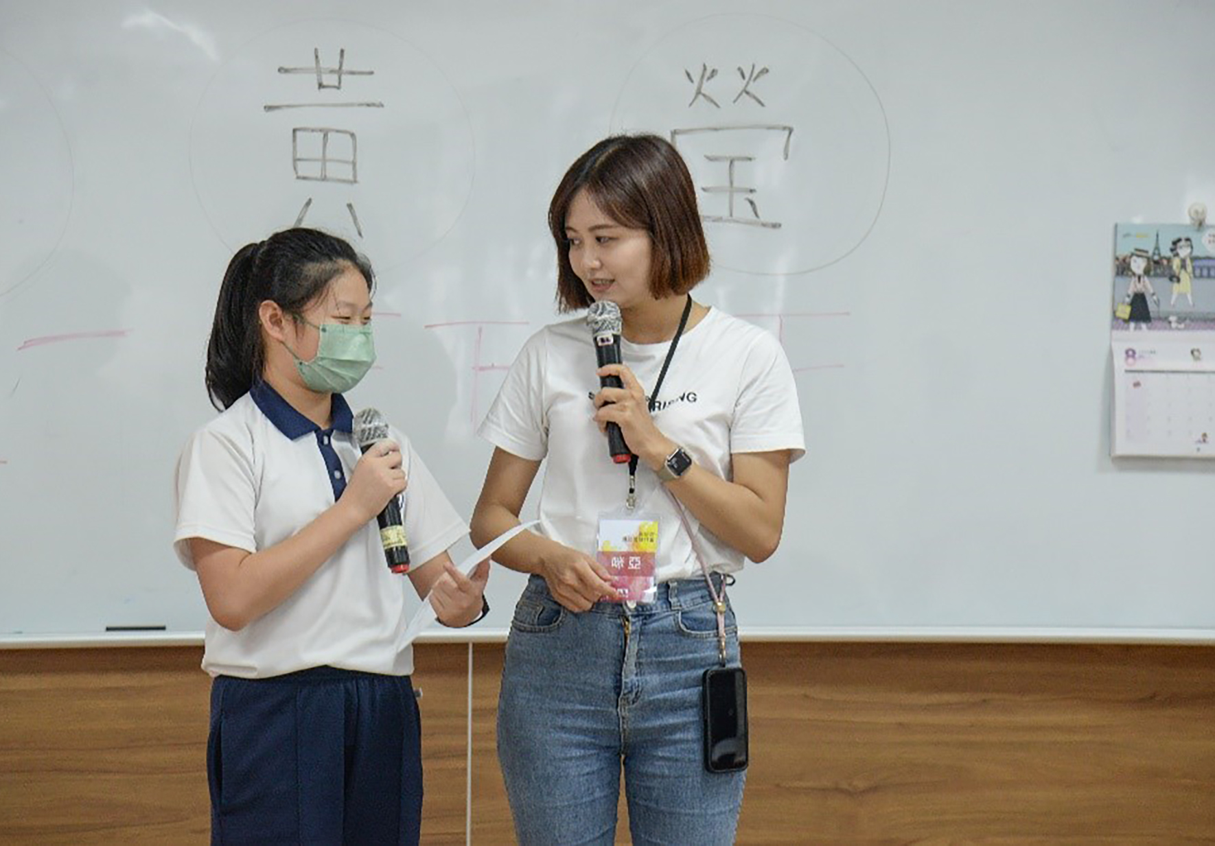 兩名女性在教室內對著麥克風進行演講。左邊穿著校服、戴著口罩的女生正在發言，右邊穿著白色上衣和牛仔褲的女生也拿著麥克風。身後白板上寫著中文字，牆上貼著活動海報。兩人都配戴識別證，呈現校園活動或教育發表會的場景。