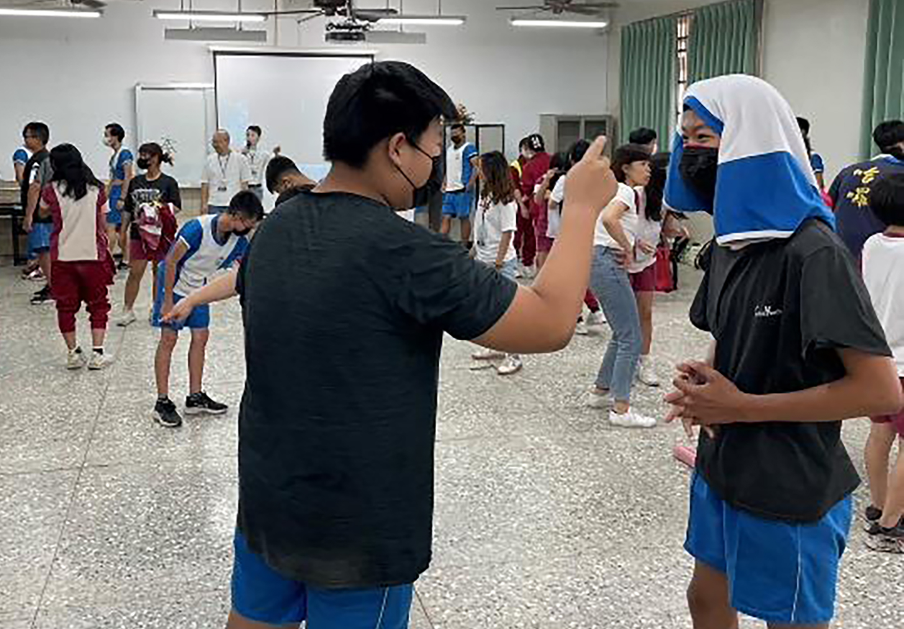 學校體育館內舉辦運動會，穿著藍色運動服的學生們在進行團體活動。前景中一名男學生與戴著藍白頭巾的女學生互動，背景有多名穿著不同隊伍服裝的學生站立，場面熱鬧。