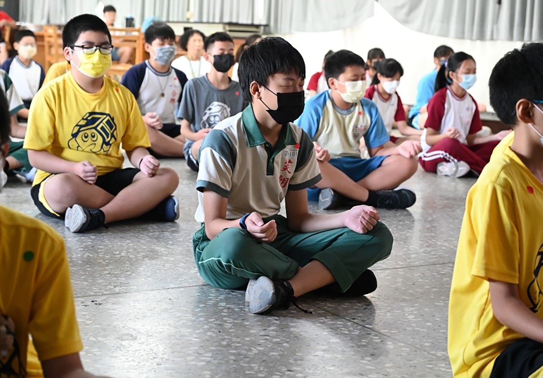 一群學生坐在室內地板上參與集體活動。多數學生穿著黃色或彩色T恤，戴著口罩。中央穿著卡其色制服的學生正帶領活動，其他學生圍坐四周專注參與。背景可見白色帳篷和木製設施。