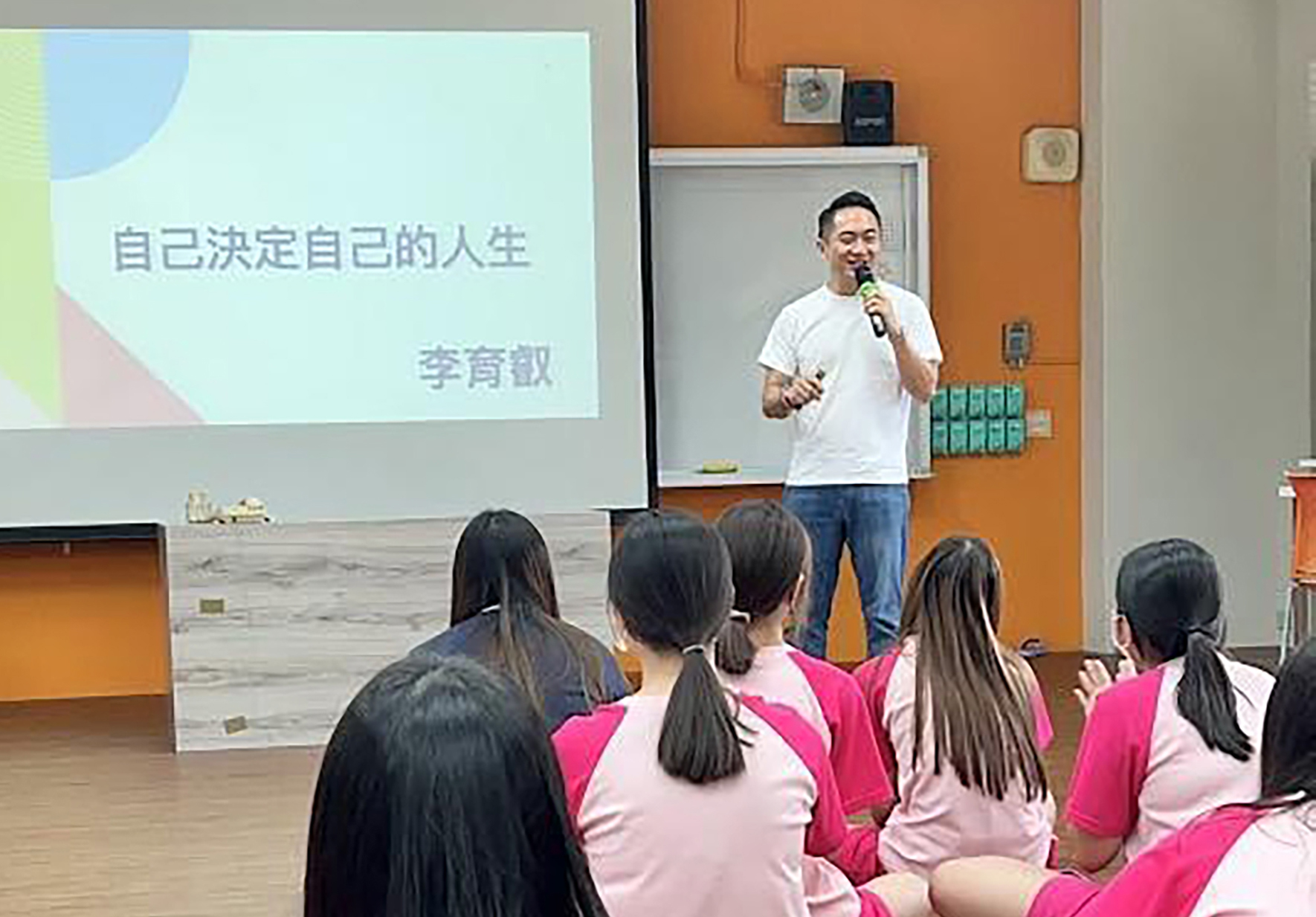 講者李育歆在教室進行演講，標題為「自己決定自己的人生」。他穿著白色T恤和牛仔褲，手持麥克風，面向坐在地上穿著粉紅色制服的學生聽眾。背景為橙色牆面和白色投影螢幕。