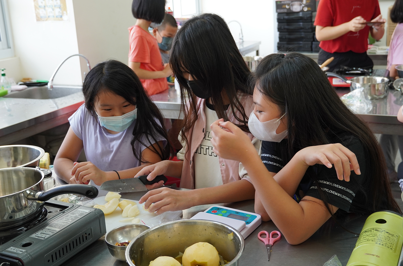 烹飪課堂中，三名戴著口罩的女學生圍在料理台前，正在切削煮熟的馬鈴薯。她們手持刀具專注於食材準備工作，身後還有其他穿著紅色衣服的學生和教師進行烹飪活動。