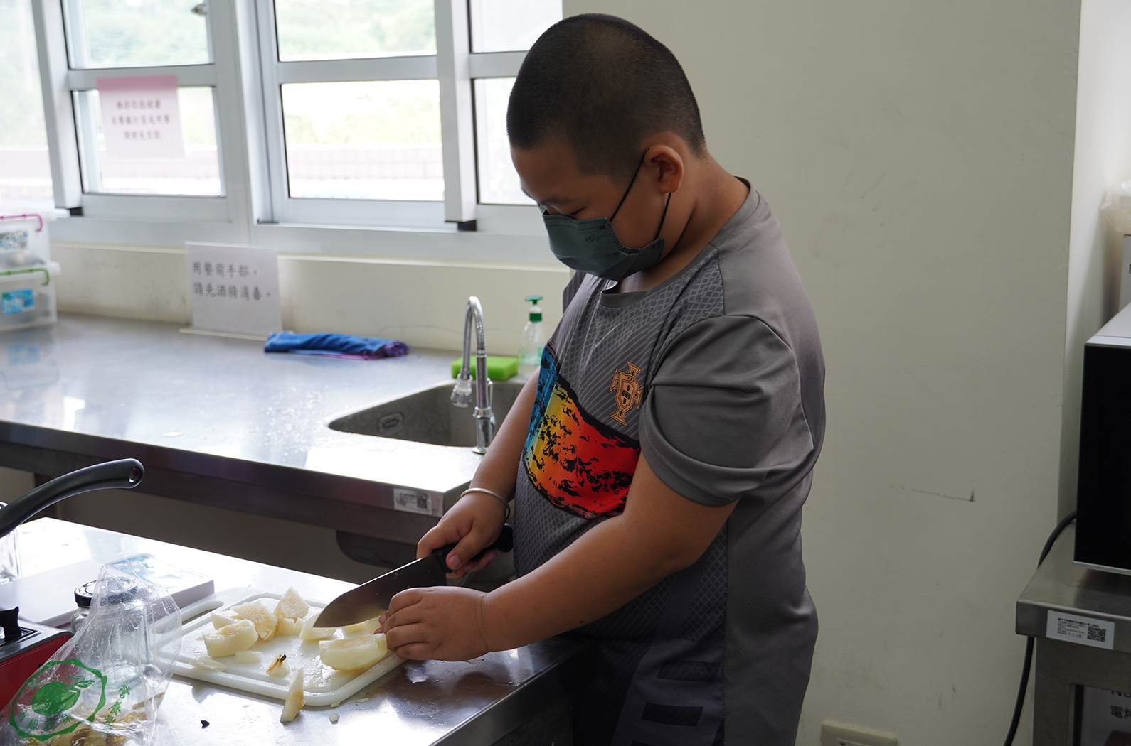 一名戴著黑色口罩的男孩在廚房實驗室的工作台上，用菜刀切割白色食材。他穿著灰色印花T恤，專注地進行食材準備工作。背景可見水槽、窗戶和廚房設備。