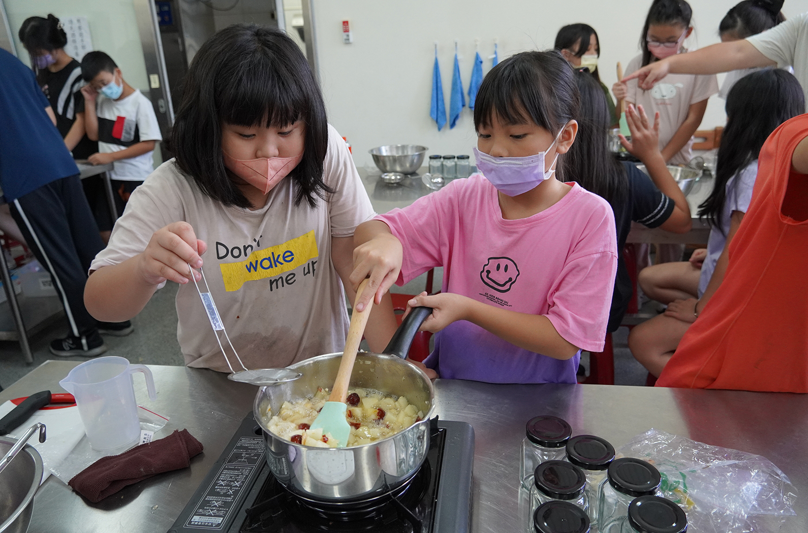 烹飪教室內，兩名戴著口罩的女學生正在爐台前合作攪拌鍋裡的食物。左邊女生穿著「Don't wake me up」T恤，右邊女生穿粉紅色T恤。背景有多名學生和老師參與烹飪活動，桌上擺放食材和調味料。