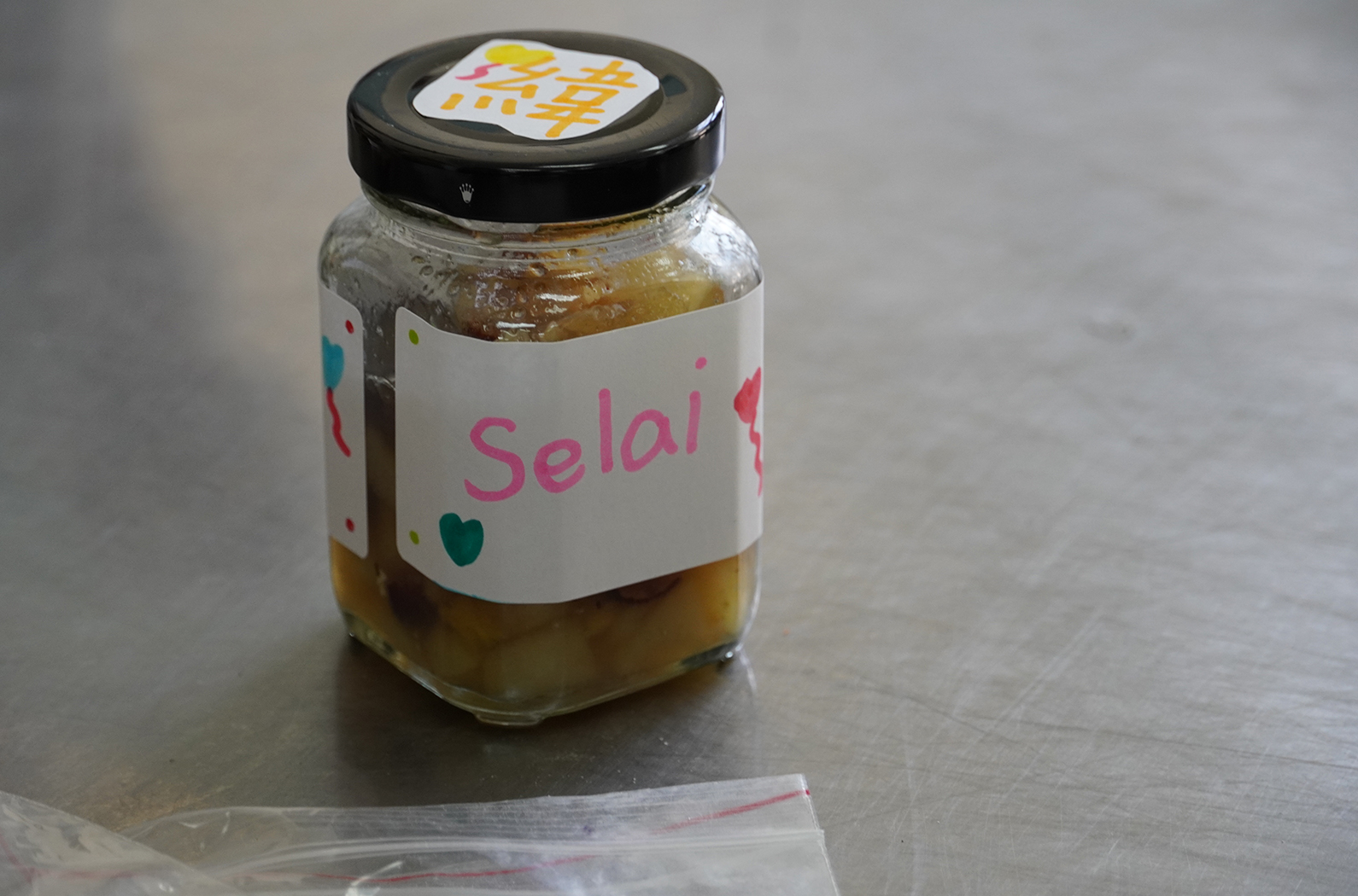 一罐玻璃瓶裝的食品，黑色瓶蓋上貼有黃色標籤。白色標籤寫著「Selai」（果醬），並附有粉紅色愛心和彩色點綴裝飾。瓶中盛裝褐色醬料。旁邊放著一個透明塑膠袋。