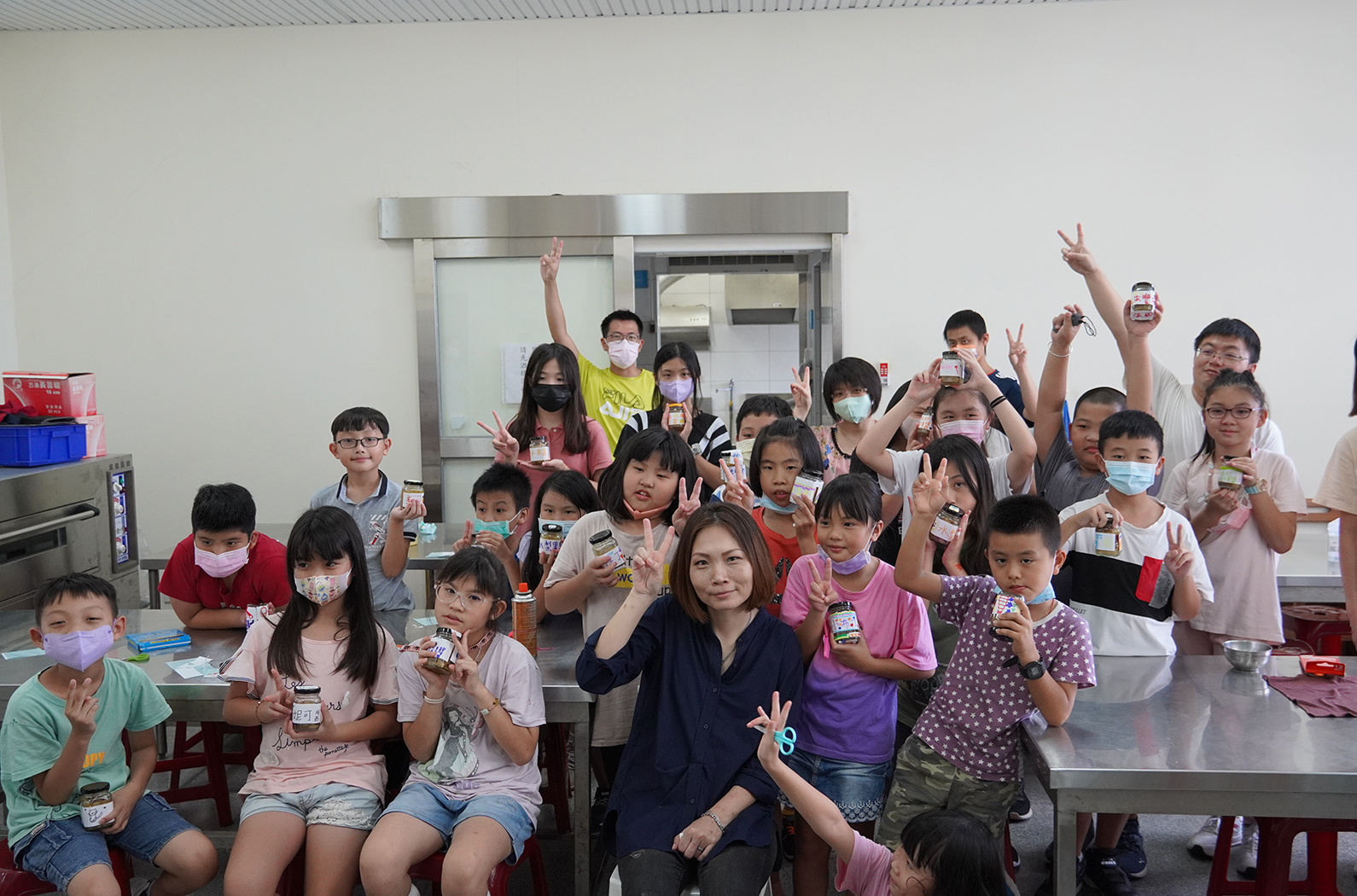一群小學生在廚房教室裡舉著食品舉起雙手慶祝，中央站著一位成年女性。大多數學生戴著口罩，展現歡樂的教學活動氛圍。