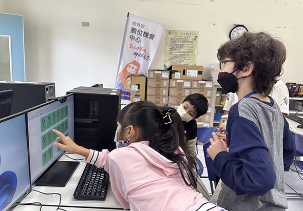 室內教室場景，三位戴口罩的學生圍繞著電腦螢幕，左側學生穿粉紅色上衣操作滑鼠，右側學生穿深藍色外套觀看。背景放置多個紙箱和宣傳海報，展示團隊協作進行電腦操作或程式設計活動。