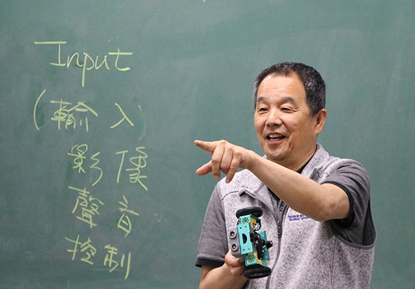一位男性教師在課堂上授課，身穿灰色短袖上衣，手持教學用具，面帶笑容指向黑板。黑板上用粉筆寫著「Input（輸入）」和中文字樣，展示教學內容。