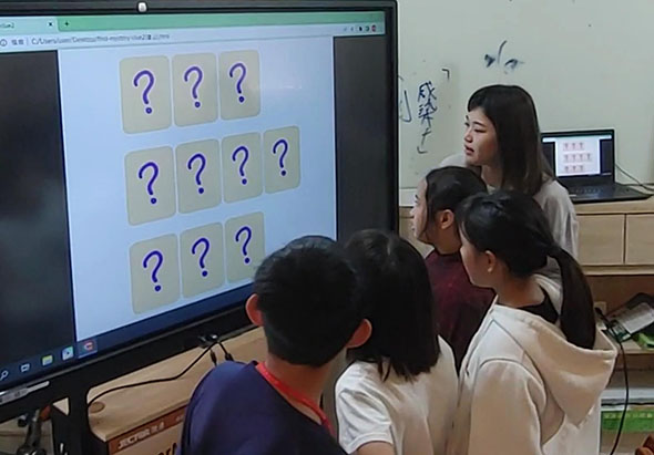 教師在課堂上使用電子螢幕進行互動教學，螢幕上顯示九個標有問號的卡片，呈現記憶配對遊戲。四位學生聚集在螢幕前觀看和參與學習活動。
