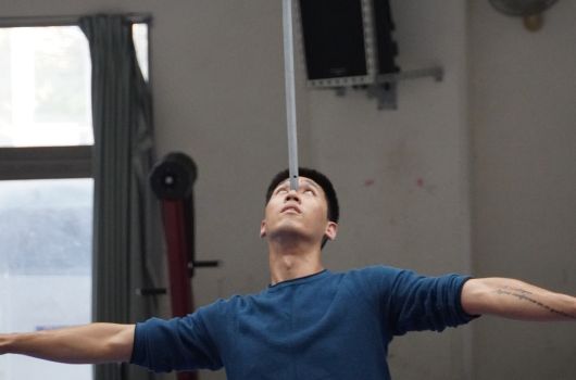一位穿著深藍色上衣的男性在室內做伸展運動，雙臂向兩側打開，頭部微微向上仰望。身後是窗戶和灰色牆面，室內光線明亮。