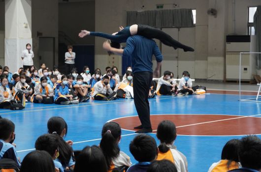 體育館內，一名男性教練在藍色運動地板上做出高難度的跳躍動作，展示武術或體操技巧。數百名穿著校服的學生坐在地板邊緣觀看表演，認真欣賞這場示範教學。