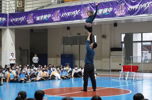 室內體育館進行籃球表演，一名男性教練在藍色地板上做跳投動作示範，身後坐著數十名穿著藍白制服的學生觀眾。場館上方懸掛紫色廣告布幡，展現專業運動教學場景。
