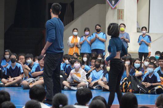 學校室內體育館舉辦活動，眾多穿著藍色制服的學生坐在地上聽講，台上有成人講師和站立的學生代表。現場防疫措施齊全，學生們配戴口罩參與集會活動。