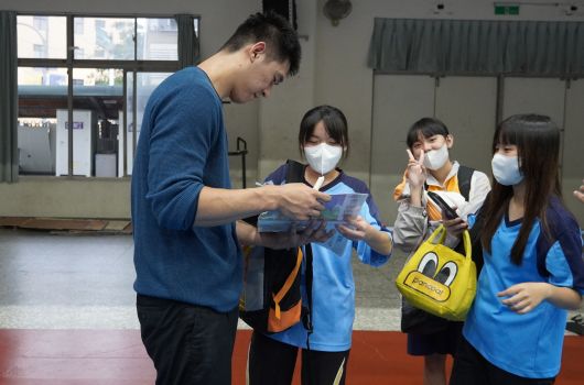 一位穿著藍色上衣的男性講師在室內體育館為四位戴著口罩、穿著藍色運動服的學生示範或講解某項活動。學生認真聆聽並觀看講師的動作演示，背景可見大型窗戶和牆壁設施。