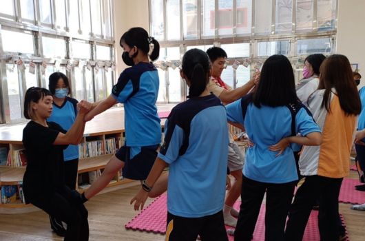 一群穿著藍色運動服的學生在明亮的教室裡進行團體活動，他們圍成一圈，雙手舉起參與互動遊戲。教室裡有書櫃和粉紅色瑜伽墊，窗戶採光良好。