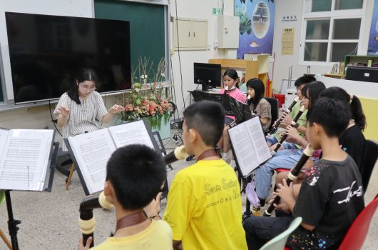 教室內，一位教師在左側指導，約十名學生圍坐成半圓形，手持樂譜進行合唱或樂器練習。教室後方有黑板、鮮花裝飾和教學用具。學生穿著各異，認真參與音樂課程。