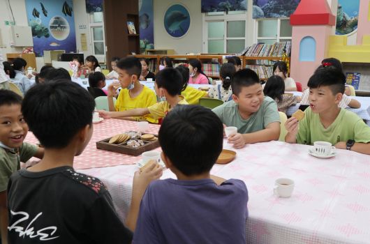 小學教室內，兒童穿著綠色和黃色制服，圍坐在粉紅色餐桌周圍進食。背景為書架、彩色儲物櫃和教室佈置。約二十多名學生在老師帶領下享用午餐或點心，呈現溫馨的校園用餐情景。