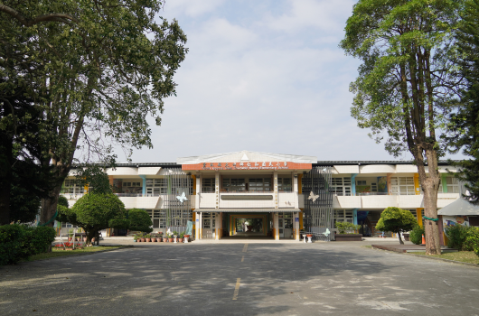 現代風格的校舍建築物，白色和棕色外觀，中央有大型入口門廊。寬闊的前廣場，兩側各有一棵高大樹木，校舍前方立有標誌牌。建築物為二層樓設計，周圍環境綠化良好。