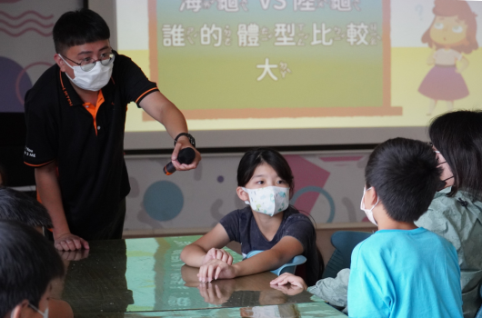 教室內，戴著口罩的講師向坐著的小學生們進行互動教學。投影幕上顯示中文課程內容，講師正用手勢與學生溝通，學生們專注聆聽。現場有多名學童參與這場教育活動。
