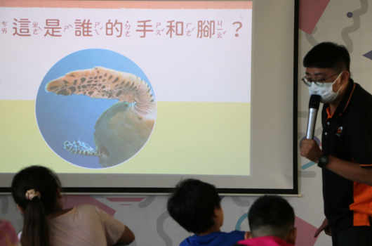 教室裡進行生物教學。投影幕顯示一隻海洋生物的圖片，上方用中文問「這是誰的手和腳？」。講師戴著口罩，持麥克風向三位學生講解。學生坐在前排專注聆聽。