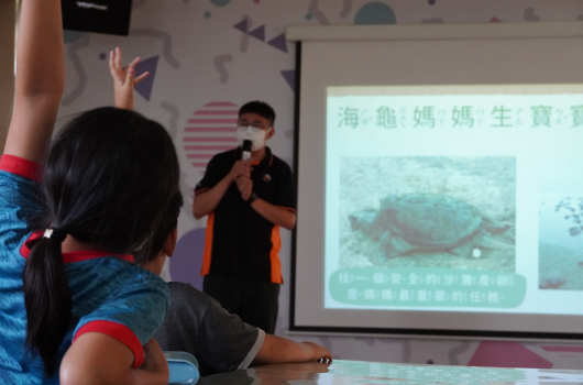 教室內進行海洋生物教育講座，講者穿著黑色上衣站在投影幕前，手持麥克風介紹海龜母親生育的相關資訊。投影畫面顯示海龜影像和中文標題。前景可見學童舉手發問，認真聆聽講解。