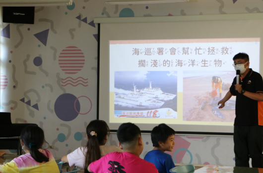 教室內，講師向學生展示關於海洋生物的投影片。螢幕上顯示海巡署巡邏船隻和海洋生物照片，搭配標題「海巡署會幫忙找、扶、救、撈淺的海洋生物」。四名學生坐在前排認真聆聽。教室牆面裝飾有幾何圖案。