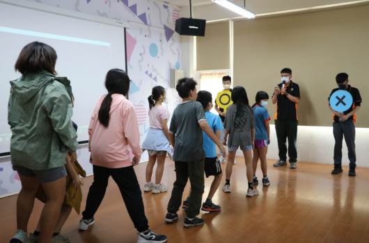 一群學生在教室內參加互動遊戲活動。學生們聚集在木質地板上，面向前方的投影螢幕。兩名老師或講師站在右側，指導活動進行。牆上貼有學習教材和圖表。學生們穿著休閒服裝，專注參與課堂活動。