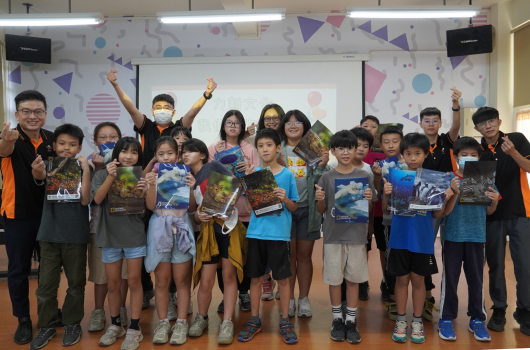 一群小學生和兩位成人在教室內舉辦慶祝活動，背景有彩色三角旗和圓點裝飾。學生們穿著各色衣服，高舉雙手歡呼慶祝，展現歡樂的班級活動氛圍。