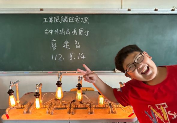 一名穿著紅色衣服、戴眼鏡的學生在教室黑板前展示自己製作的橙色科學實驗裝置，裝置上裝有多個發光燈泡和金屬結構，學生做出勝利手勢，黑板上寫有中文課題和日期。