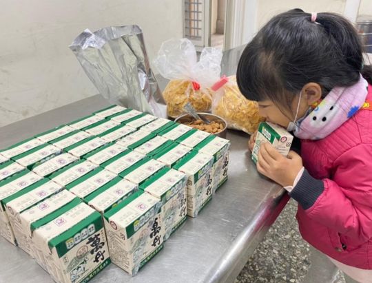一名戴著粉紅色口罩的女性在檢查整齊排列的白色和綠色紙箱。桌面上擺放著多排紙箱，旁邊還有塑膠袋裝的食品和其他物品，背景是室內空間。