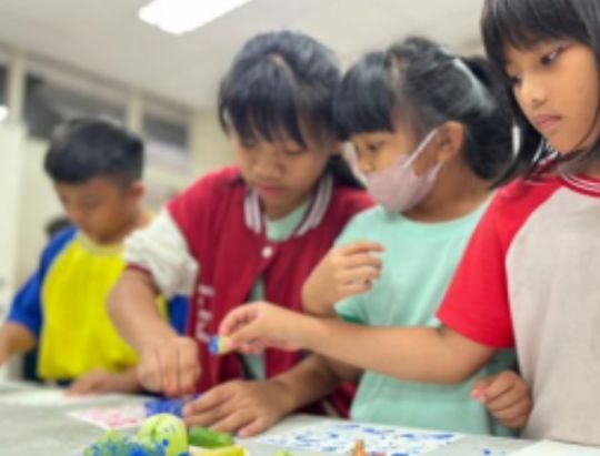 一群小學生在教室內圍聚一起進行美術創作活動，穿著紅色和彩色衣服的孩子們正專注地在紙張上畫畫和著色，其中一位學生戴著口罩，展現出小學生們認真參與創意藝術課程的情景。
