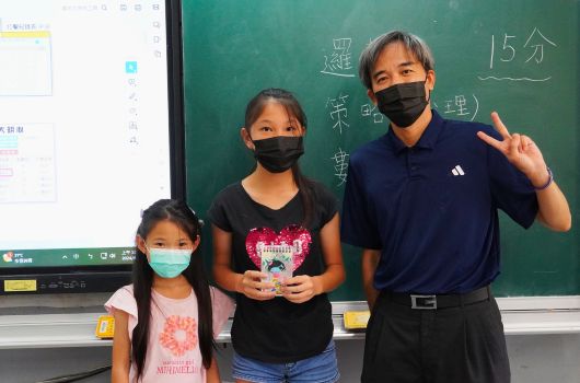 教室內，一名成年男性與兩名女童站在綠色黑板前合影。三人均戴著黑色口罩。黑板上有中文字跡，身後可見電子互動白板。男性穿著深藍色polo衫，做出手勢。這是一張師生互動的校園照片。