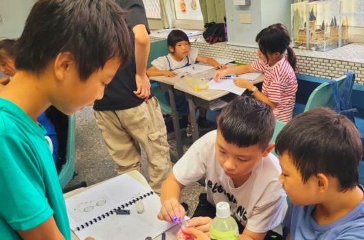 教室內多名小學生圍繞著桌子進行美術創意活動，學生們正在用彩色紙張和材料進行手工製作，展現專注和互動的學習氛圍。教室背景可見教學用掛圖和學習資源。
