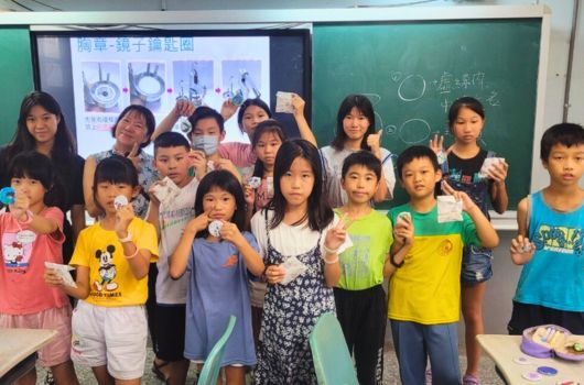 教室裡約二十位小學生穿著各色衣服，做出勝利手勢面向鏡頭。身後黑板上寫有中文字，投影幕顯示眼睛相關圖片。這是一場學校視力保健教育活動的合照。