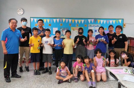 教室內一群小學生和一位成人教師排成一列，做出拳頭手勢。教室背景有藍色三角旗裝飾和布告欄。學生穿著各色上衣，有人坐在前排，大部分站立。牆上掛著時鐘。這是一場教育活動或班級合照。