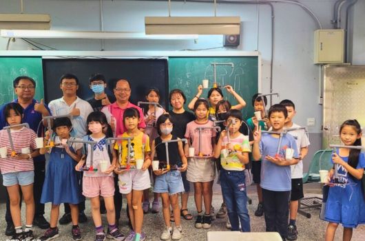 教室內，教師與約二十名小學生站在黑板前合照。學生們穿著各色夏季校服，開心地展示手中的獎狀或證書。黑板上有粉筆畫的圖案。這是一場校內頒獎或表揚活動的集體合影。