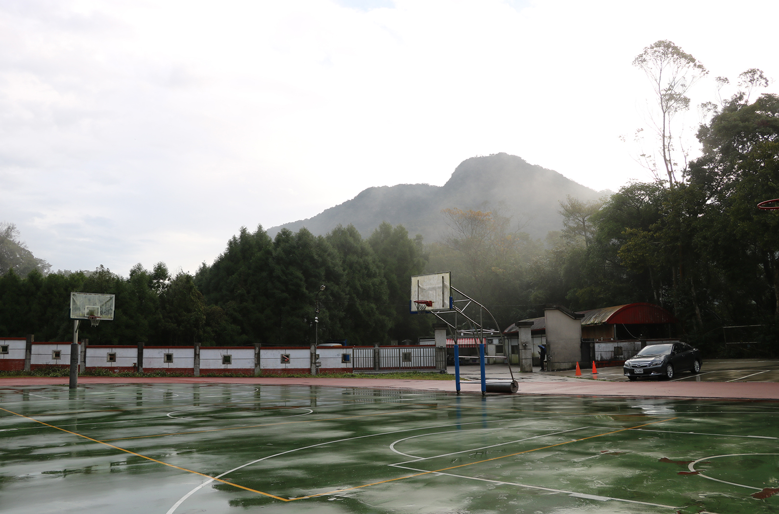 雨後的籃球場，濕潤的地面反射著天空。左側是籃球架，中央和右側各有一個籃框。背景是蒼翠的樹林和籠罩在霧氣中的山峰。場邊有建築物和停放的黑色轎車。