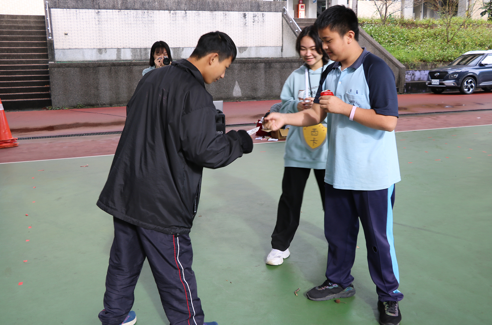 四名學生在室外網球場進行團隊活動。穿著黑色外套的學生正在向其他三位同學展示或分發某個物品。背景可見停車場、建築物和綠色植被。地面為綠色和紅色的運動場地。