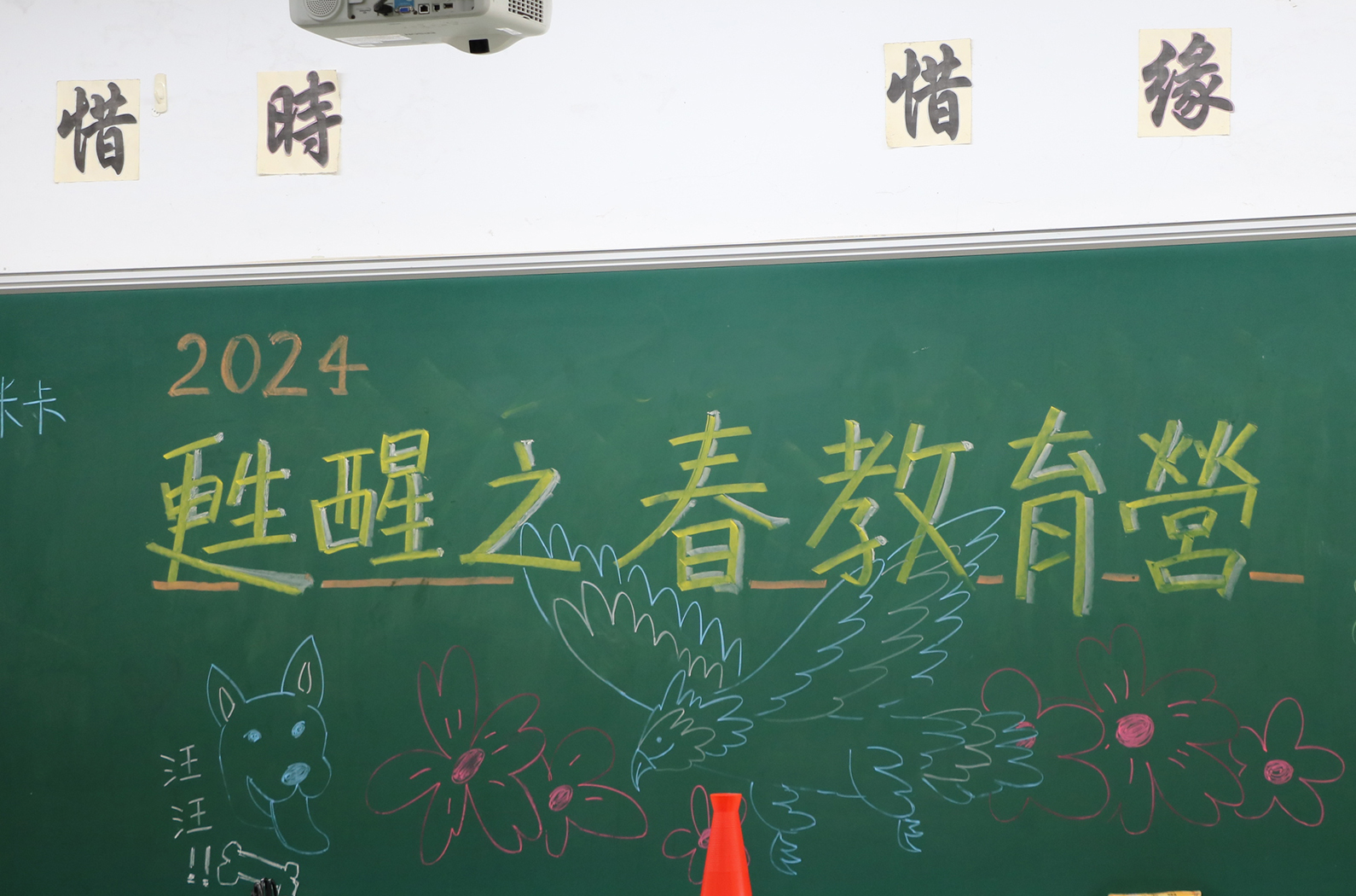 教室黑板上書寫2024年題目「躍躍之春教育營」，用黃色粉筆寫成。黑板下方繪有彩色的動物和花卉插圖，包括兔子、蝴蝶和花朵。黑板上方貼有四張黃色標簽，寫有中文字。教室天花板安裝有投影機。