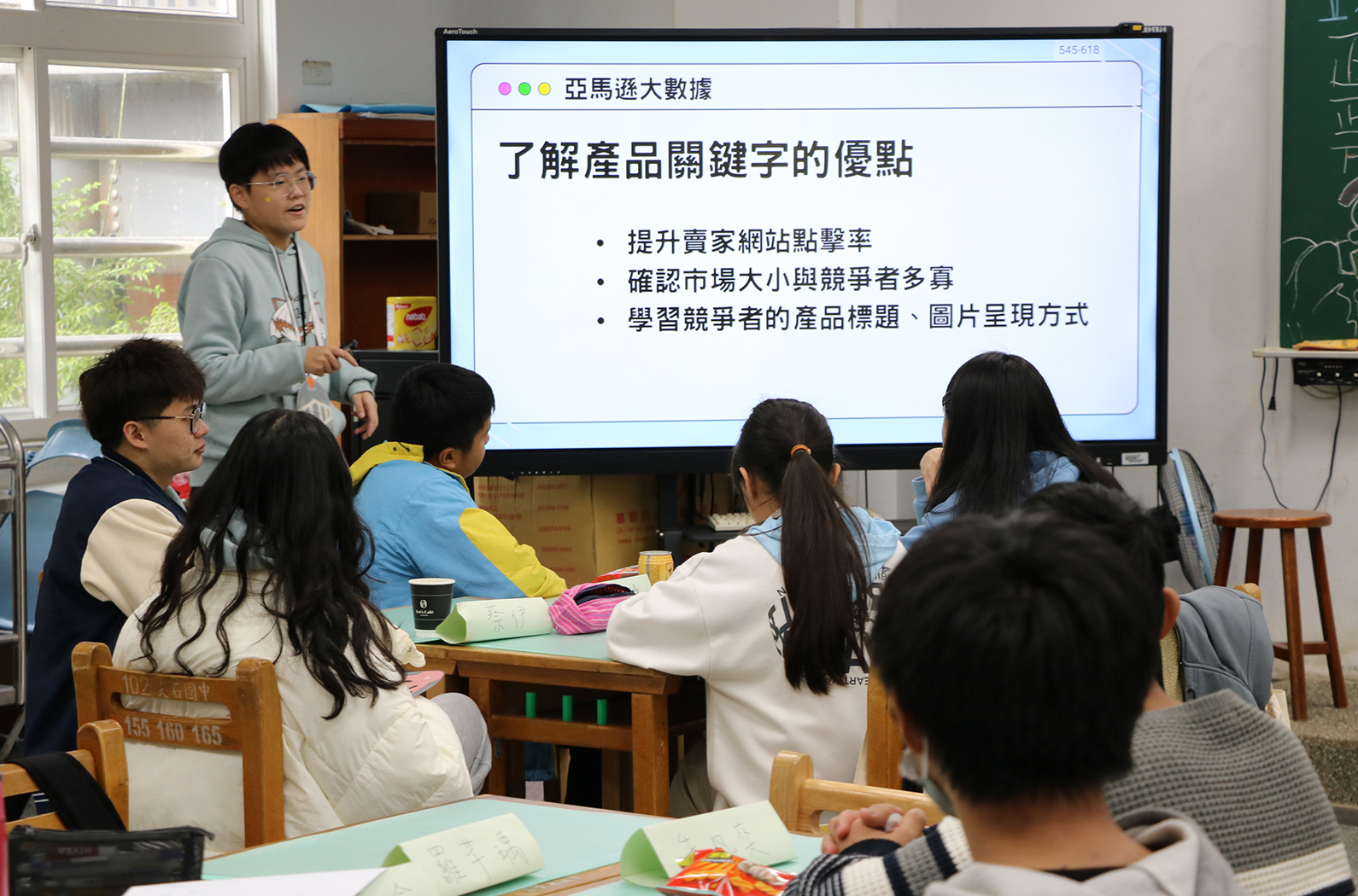 教室內，一位講師站在投影螢幕前向學生講授「亞馬遜大數據」課程。螢幕上展示「了解產品關鍵字的優點」主題，列舉提升賣家網站點擊率、確認市場大小與競爭者、學習競爭者的產品標題和圖片呈現方式等三點。約六位學生圍坐在木製桌前認真聽講。
