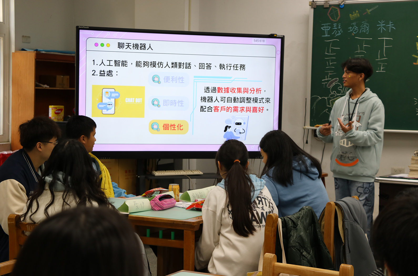 教室內，一位講師向五名學生介紹「聊天機器人」主題。大螢幕展示簡報，說明人工智能的功能與益處，包括便利性、即時性和個性化。學生坐在木製課桌前專注聆聽，黑板上有中文字跡，教室角落放有木製櫃子。