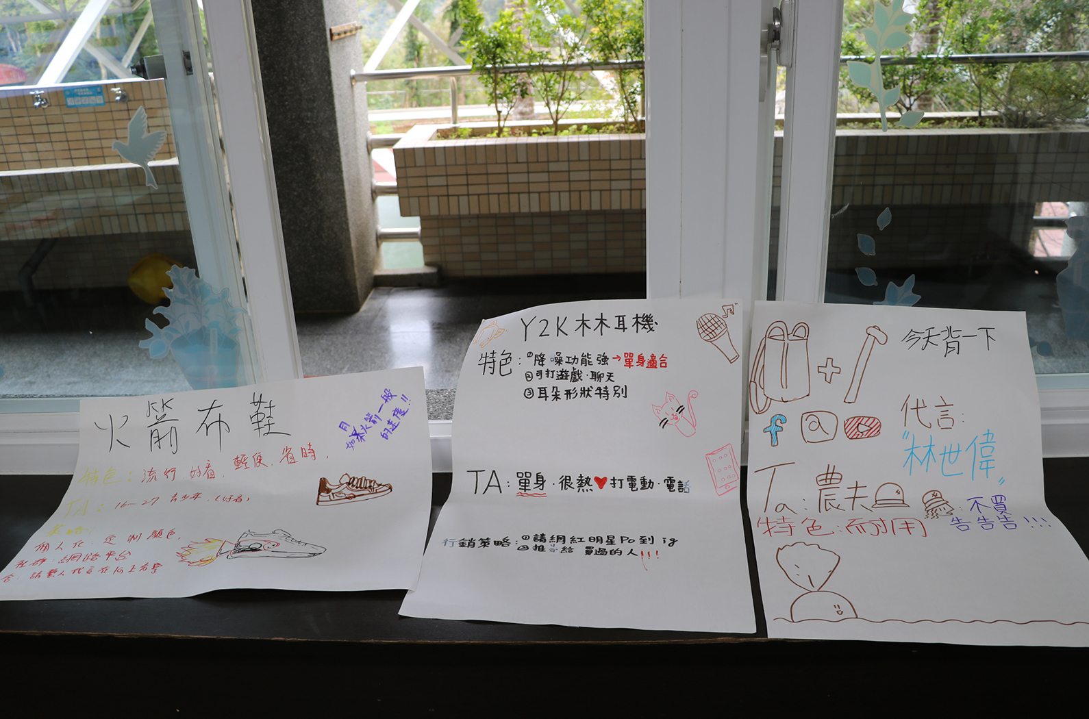 窗邊展示三張手繪海報，分別介紹運動鞋、Y2K木木耳樓和領帶等主題。每張海報用彩色筆繪製，包含文字說明和簡單圖案。背景可見建築物和綠色植物，呈現學生作品展示的場景。