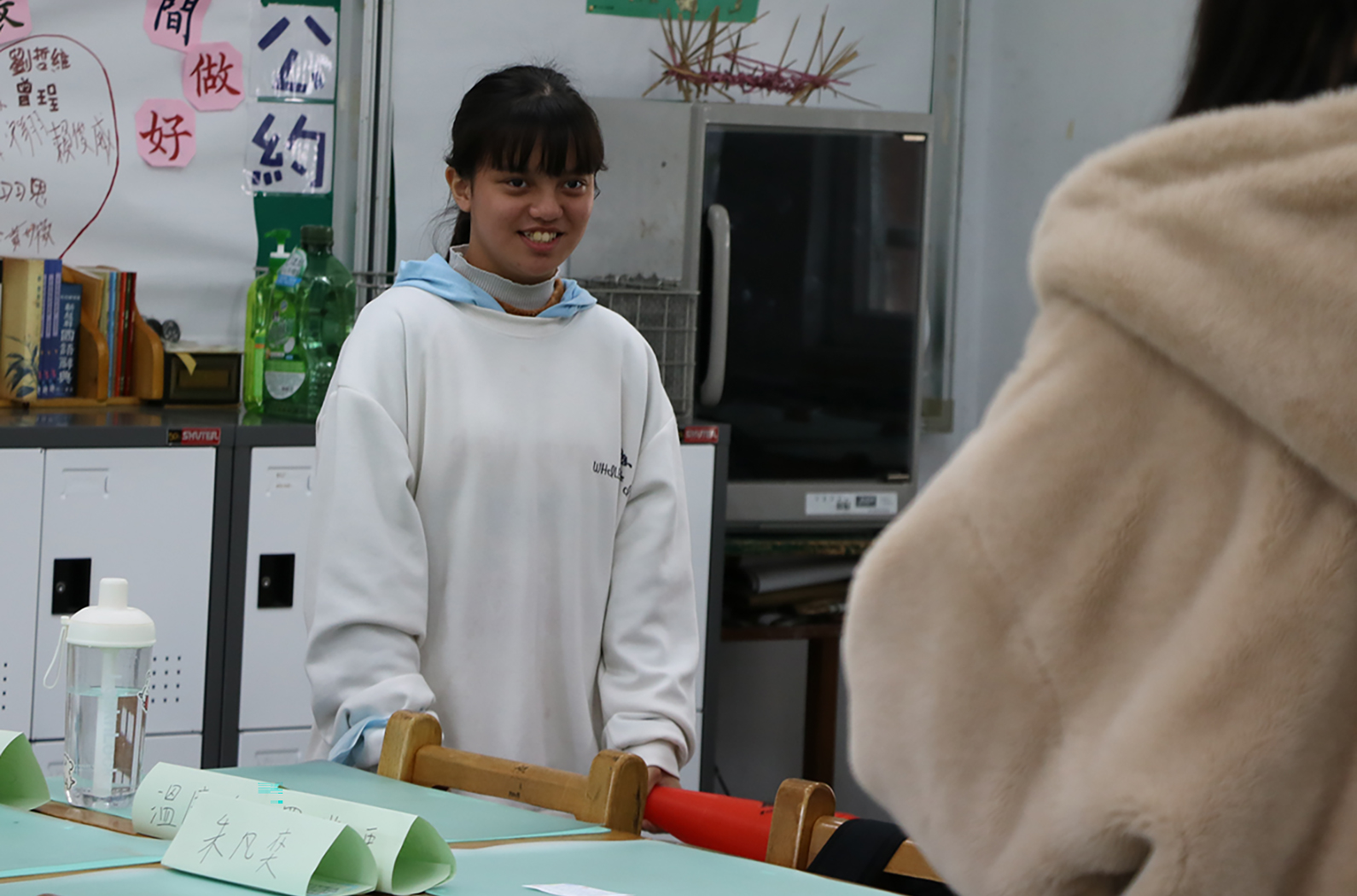 教室內，一位穿著白色連帽衫的女學生站在講臺後方，面帶微笑。背景的白板上貼有中文教學卡片，講臺上放置著教學用具，包括綠色三角形紙製品和紅色筆筒。教室設有冰箱和其他教學設備。