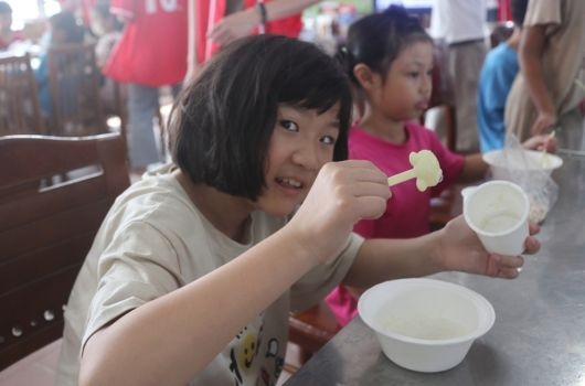 一名穿著淺色T恤的女孩在室內活動現場享用食物，手持白色餐具和杯子，面帶笑容。身後有其他參與活動的人員，背景可見紅色布幔和其他參加者。